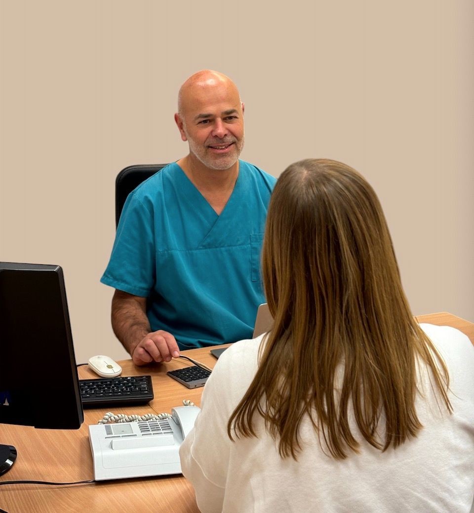 Doctor in blue scrubs smiling at a woman sitting across a desk in an office setting.